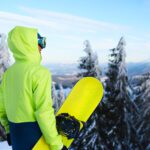 Back view of snowboarder standing with his board on the mount before backcountry freeride session in the forest showing what to pack for Big Sky Montana